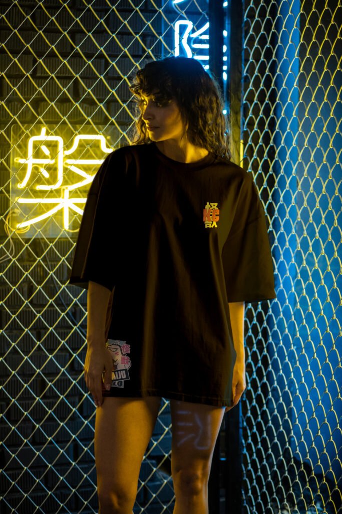 Modelo feminina vestindo camiseta preta oversized da Kaiju Clo em um cenário urbano noturno com luzes de neon japonesas ao fundo.